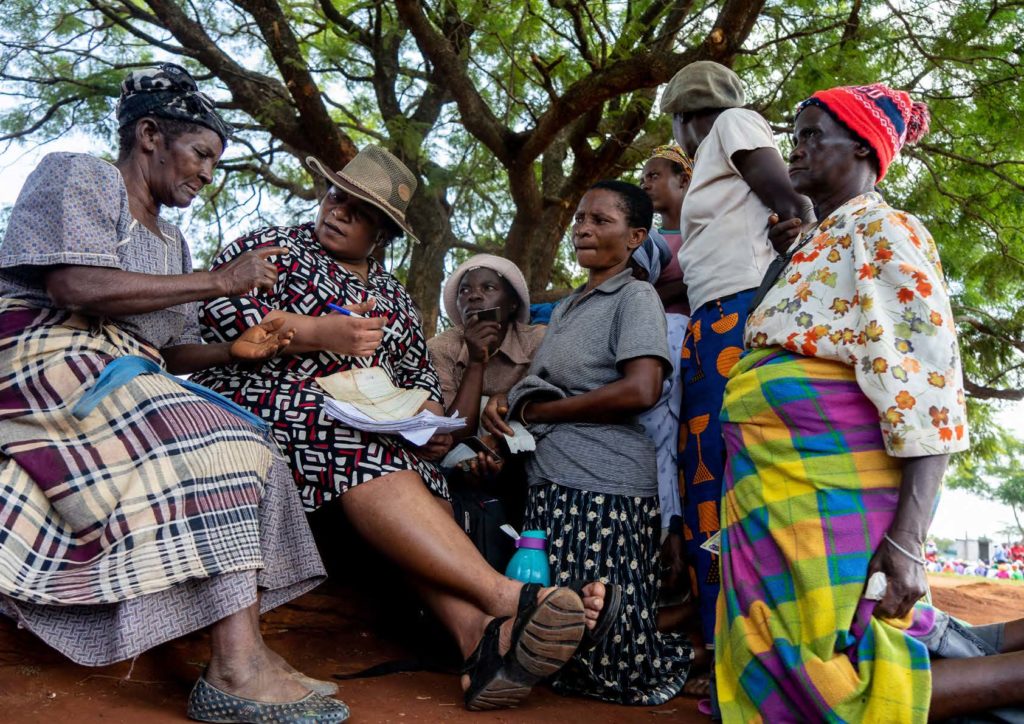 Group of women wearing floral and African print in discussion outdoors under a tree, one woman holds a pen and pad of paper