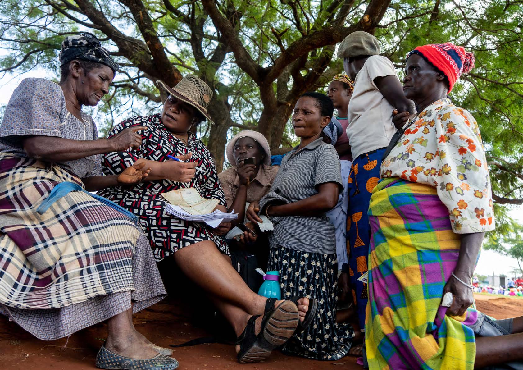Group Of Women Wearing Floral And African Print In Discussion Outdoors Under A Tree, One Woman Holds A Pen And Pad Of Paper