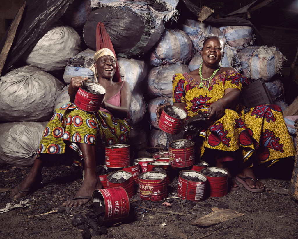 Two black women wearing African print selling cans of charcoal sat in front of large bags of charcoal in Togo