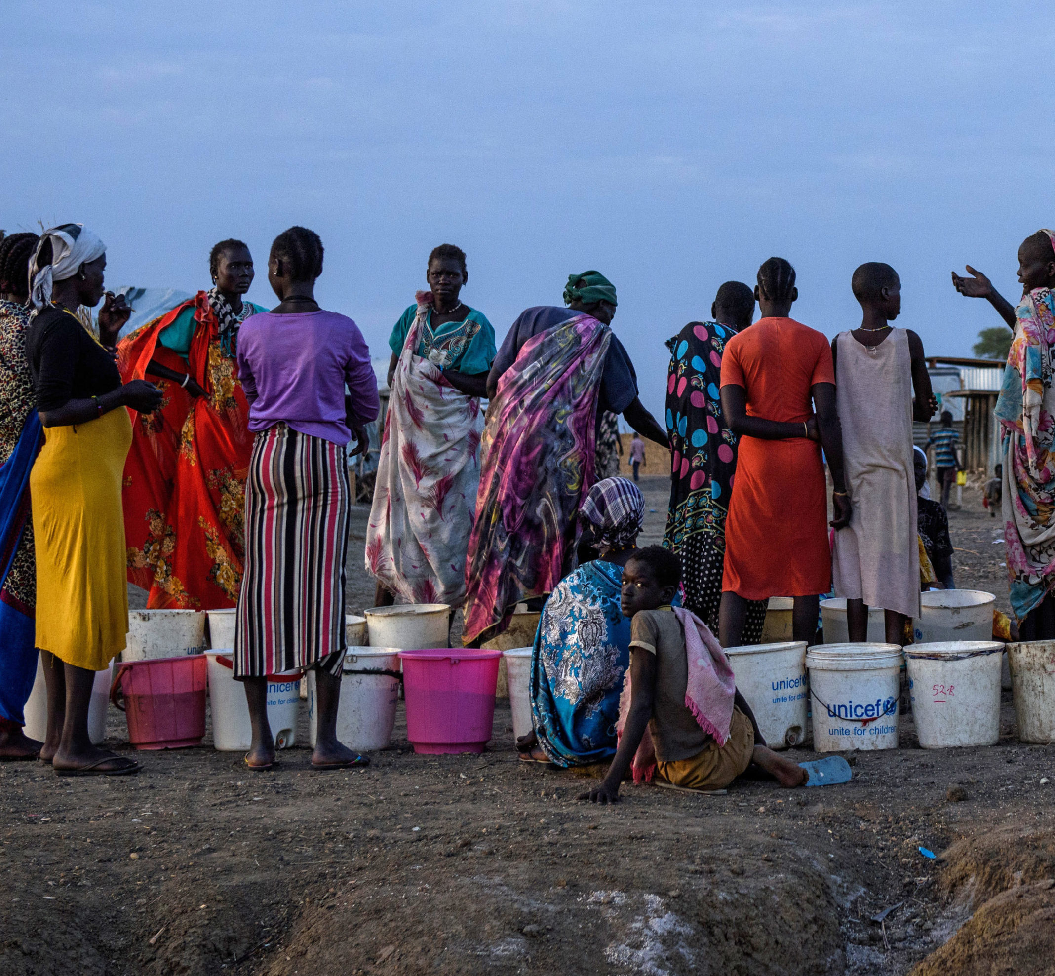 African Women Standing And Talking With UNICEF Labelled Buckets At Their Feet, One Woman And Boy Are Sitting, The Boy Is Looking At The Camera