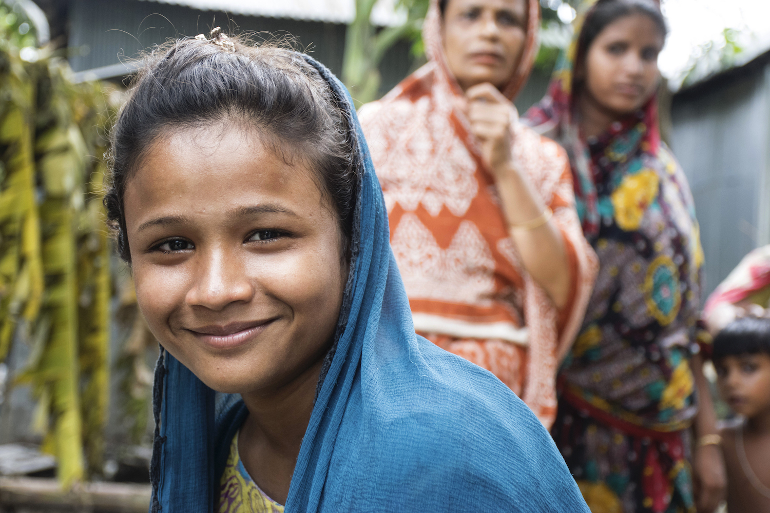 Bangladeshi Girl Wearing A Headscarf Smiling At Camera, Two Women And A Boy Stand Behind Her