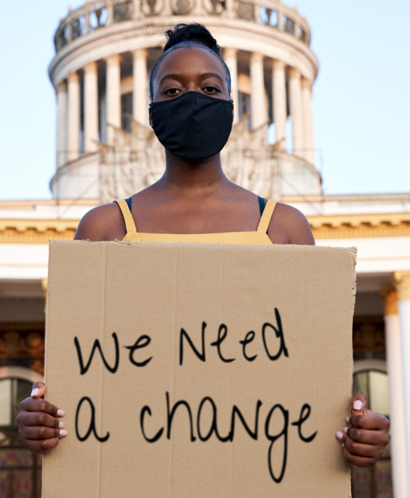Black woman wearing mouth mask holding a placard saying we need a change