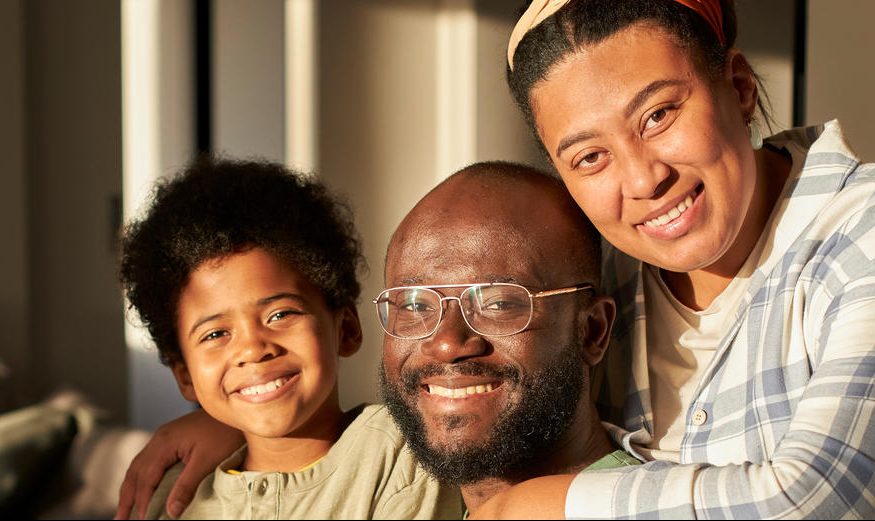 A Mixed Race Family Of Three, A Women, Man And Child Smiling And Hugging.
